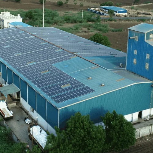 An aerial view showcases a large industrial building with a blue roof adorned with Surya Shakti solar panels, efficiently harnessing energy while surrounded by lush greenery.